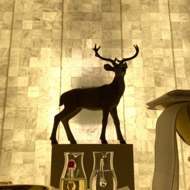 Hirschfigur auf Sockel, davor ein Altar mit Wasser und Wein, Hostienschale und Kelch. Die Wand im Hintergrund erstrahlt in goldenem Licht.