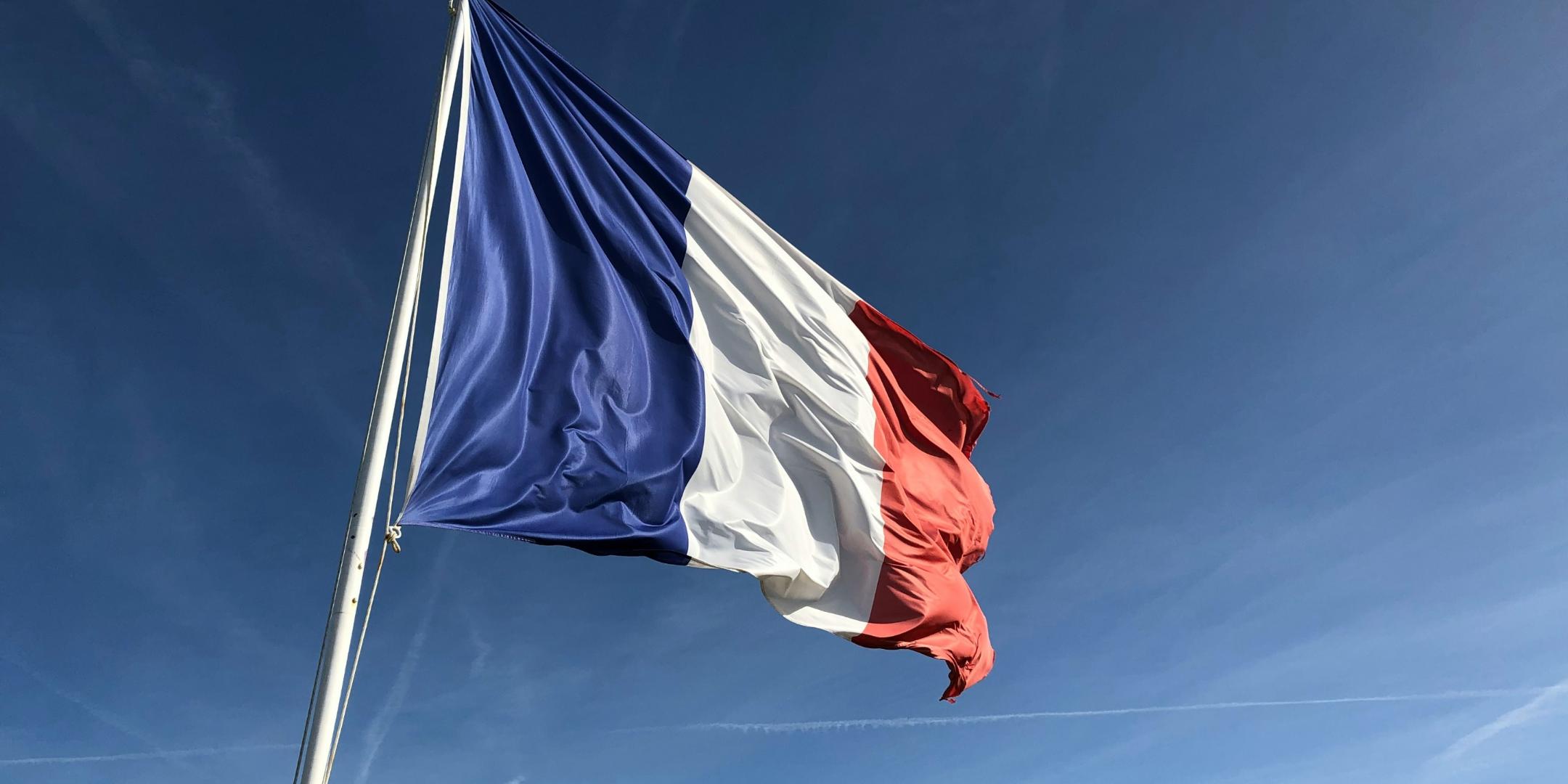 Auf dem Bild ist die französische Flagge in den Farben blau, weiß und rot vor einem blauen Himmel zu sehen (c) Foto von Anthony Choren auf Unsplash Auf dem Bild ist die französische Flagge in den Farben blau, weiß und rot vor einem blauen Himmel zu sehen
