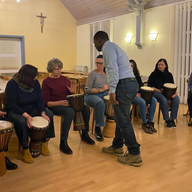 Eine Gruppe von Menschen sitzt in einem hell erleuchteten Raum in einem Halbkreis, jeweils mit einer Djembe‑Trommel vor sich. Ein stehender Anleiter in der Mitte gibt Hinweise oder führt etwas vor. Der Raum hat helle Holzböden, hohe Fenster mit Lamellen und warmgelbe Wände mit Wandlampen. Alle wirken konzentriert und in eine gemeinschaftliche Trommelübung vertieft.