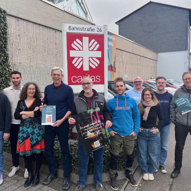 Eine Gruppe von unterschiedlichen Leuten steht draußen vor dem Caritas-Logo und schaut in die Kamera.